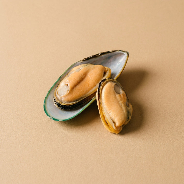 Grönläppad mussla öppnad på halva skalet med sitt aprikosfärgade kött, bredvid en urtagen mussla, på varmbeige bakgrund.